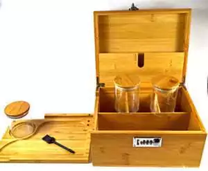 Wooden hinged storage box with a lock, open to reveal two glass jars with bamboo lids, a small wooden spoon, and an interior tray for organizing herbs and ritual supplies.