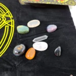 Variety of polished gemstones in different shapes and colors arranged on a black surface with a yellow circular pentagram design from the RWS tarot kit.