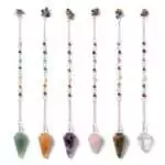 Row of six gemstone pendulums in different chakra-aligned crystal colors, each on a metal chain, chakra stone dowsing and divination tools displayed together.
