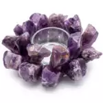 A circular arrangement of raw amethyst crystals surrounds a clear glass candle holder. The crystals are various shades of purple, creating a decorative and natural look.