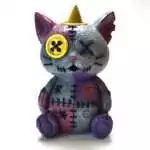 Painted resin figurine of a patchwork stitched cat with a yellow button eye, purple and gray color-blocked patches, and a yellow backflow incense cone set atop its head.