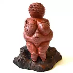 Venus of Willendorf replica statue: a red-brown figure with a banded head, rounded body, and arms across the breasts, standing on a dark rock-textured base.