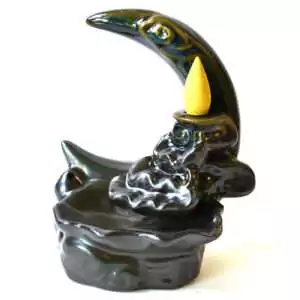 Black glazed ceramic crescent moon backflow incense burner with a sculpted eye, a yellow cone in the cone dish, and sculpted clouds above a wavy basin, on white